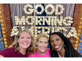 Hayley Knollman, Kate Korson, and Lola Fayanju smile in front of bright lights spelling Good Morning America.